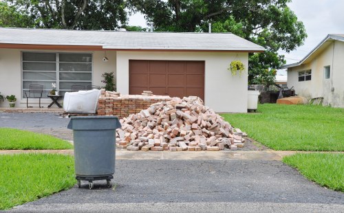 A residential driveway in Yiewsley, with a pile of red bricks and rubble placed directly on the paved surface in front of a single-story house. The house features a large window with horizontal blinds and a small potted plant on the window sill, with a brown garage door situated centrally on the front facade. To the left of the brick pile, a dark grey wheelie bin is positioned on the driveway. The surrounding area includes a well-maintained lawn on either side of the driveway, with green grass extending along the front of the property and some mature trees visible behind the house. The sky is overcast, providing diffused natural light that highlights the textures of the brick debris, the smooth finish of the garage door, and the lush greenery. This scene represents a typical outdoor setting where a rubbish removal service, such as those offered by Office Clearance Yiewsley, might be involved in clearing building waste or debris from a residential property in the local area.