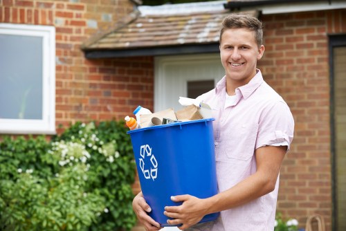A young man with short, light brown hair and a friendly smile is standing outdoors in front of a red brick house with a white-framed window and a tiled roof. He is wearing a light pink, short-sleeved button-up shirt and is holding a blue plastic recycling bin filled with assorted rubbish, including cardboard, plastic bottles, and paper waste. The bin features a white recycling symbol on the side. Surrounding him are some green shrubbery and flowering plants, indicating a well-maintained garden area. The scene suggests a residential setting during daytime, possibly after clearing waste or preparing for rubbish collection, aligning with professional waste removal services such as those provided by Office Clearance Yiewsley near the local postcode area. The image captures a moment of responsible rubbish disposal in a typical suburban environment, emphasizing the importance of proper waste management and recycling in the community.