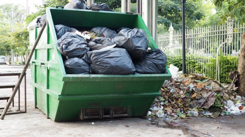 A large green skip bin filled with black rubbish bags is positioned outdoors on a paved area, surrounded by scattered litter and debris. The skip is situated near a metal fence, with trees and greenery visible in the background, indicating an outdoor space possibly within a residential or commercial area in Yiewsley. The skip's metal construction has a smooth finish with horizontal ribbing on the sides and is elevated slightly off the ground with black support blocks underneath. To the right of the skip, a pile of loose rubbish, including cardboard, paper, and other waste materials, is seen spilling onto the pavement. The environment appears to be a typical scene for rubbish collection or clearance, aligning with the services provided by Office Clearance Yiewsley for local rubbish removal and waste management.