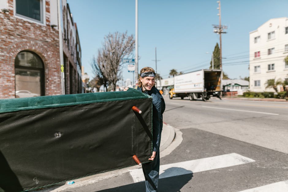 A street scene in an urban area shows a large white waste collection vehicle parked parallel to a curb, with its rear open revealing rusted mechanical components and a partially concealed waste hopper